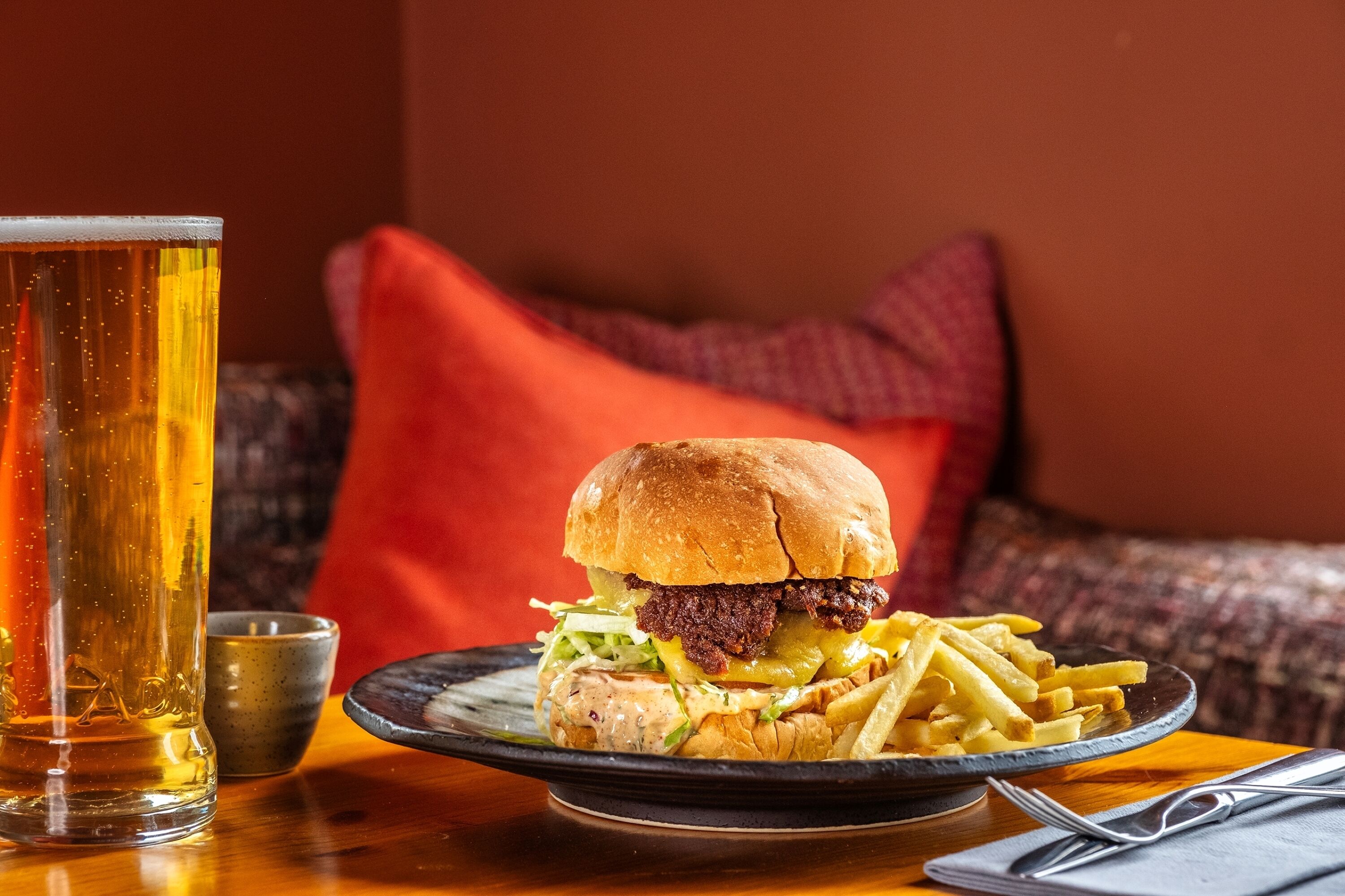 A meal with a burger and fries on a plate, a glass of beer, and a small cup on a table.