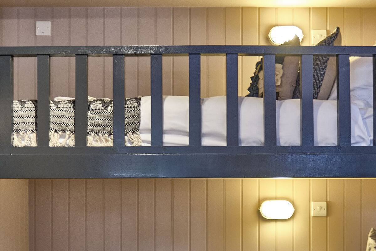Een stapelbed met een beddengoed met patroon en kussens, een onderbed met beddengoed en kussens, en een wandlamp.