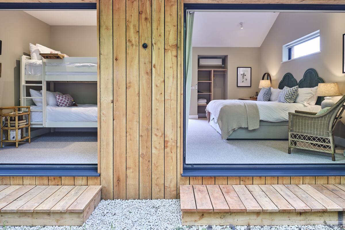 Een kamer met stapelbedden, een bed met gestreepte kussens, een rieten stoel en een raam met uitzicht.