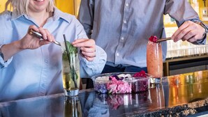 Baaritiski, jossa baarimikko valmistaa juomaa, asiakas pitää kädessään cocktail-lasia ja hyllyt ovat täynnä erilaisia alkoholijuomapulloja.