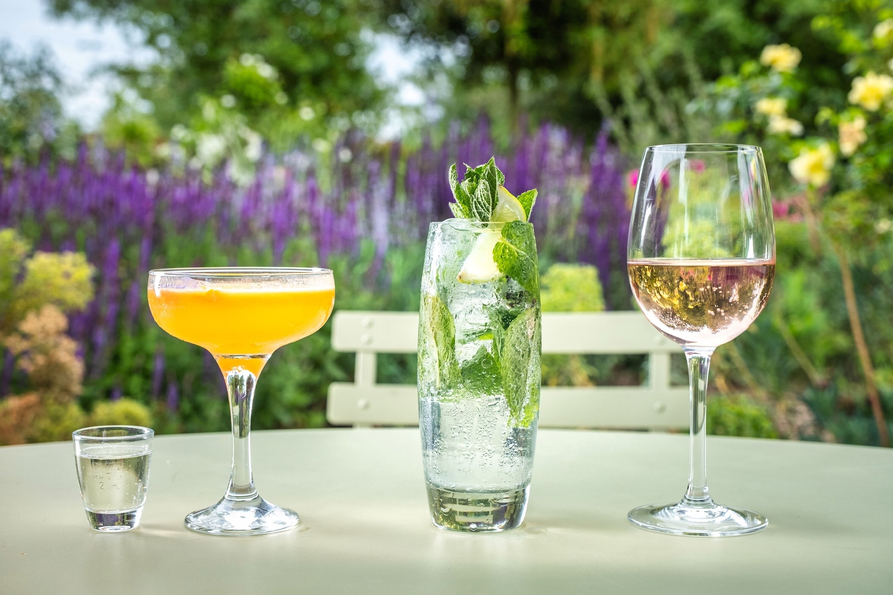 Three glasses of beverages on a table with a white bench and purple flowers in the background.