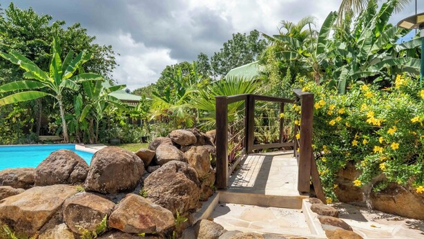 Un sendero de piedra que lleva a una pérgola de madera rodeada de plantas tropicales y una piscina.