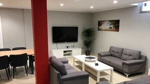A modern living room with a flat-screen TV, grey sofas, a white coffee table, and a dining area with a wooden table and black chairs.