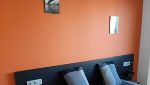 A bedroom with an orange wall, a bed with a checkered bedspread, two pillows, a nightstand, and a small shelf.