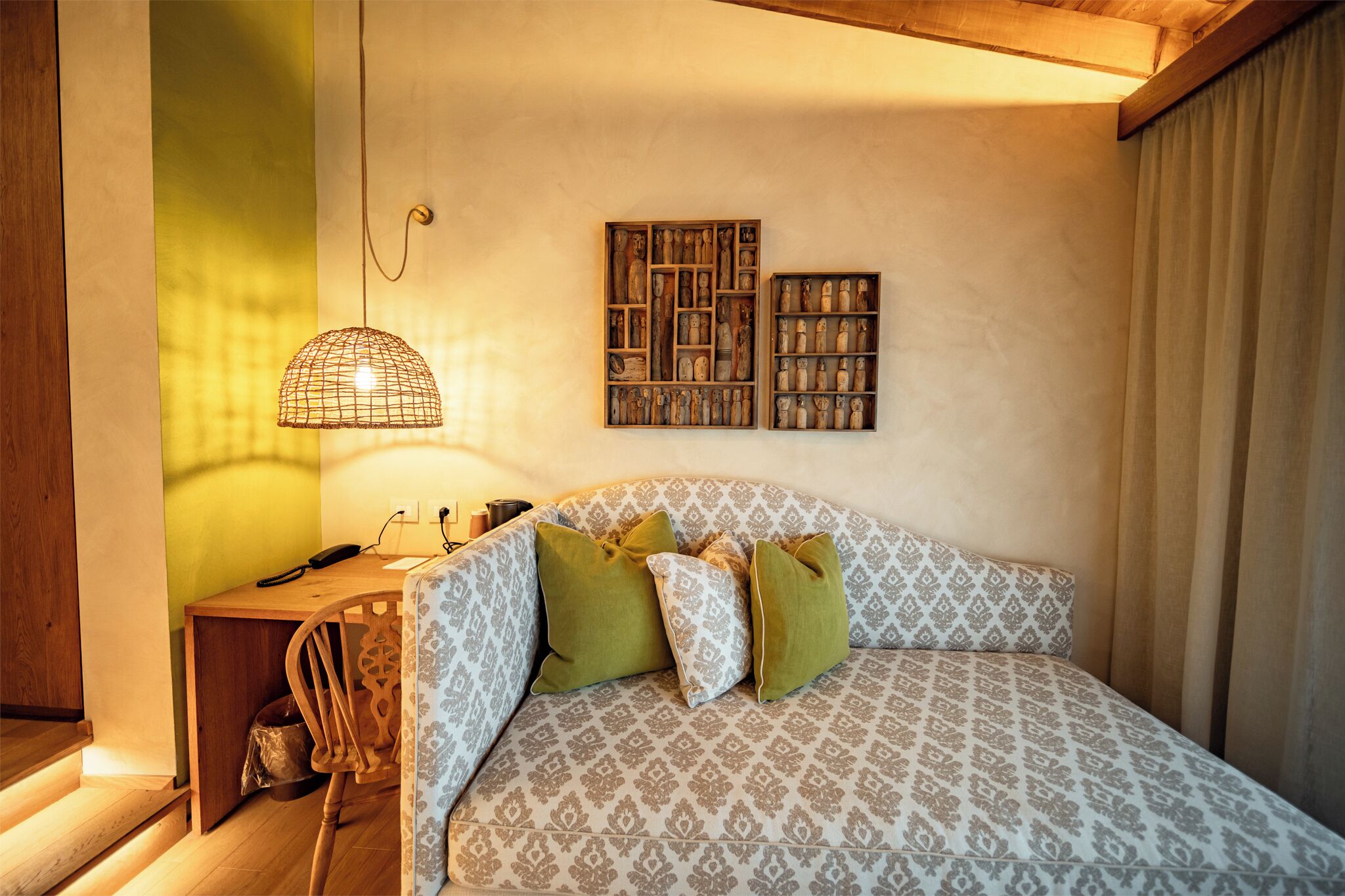 Une chambre à coucher avec une couvre-lit à motifs, un bureau en bois avec une lampe et deux étagères murales ornées d’objets décoratifs.
