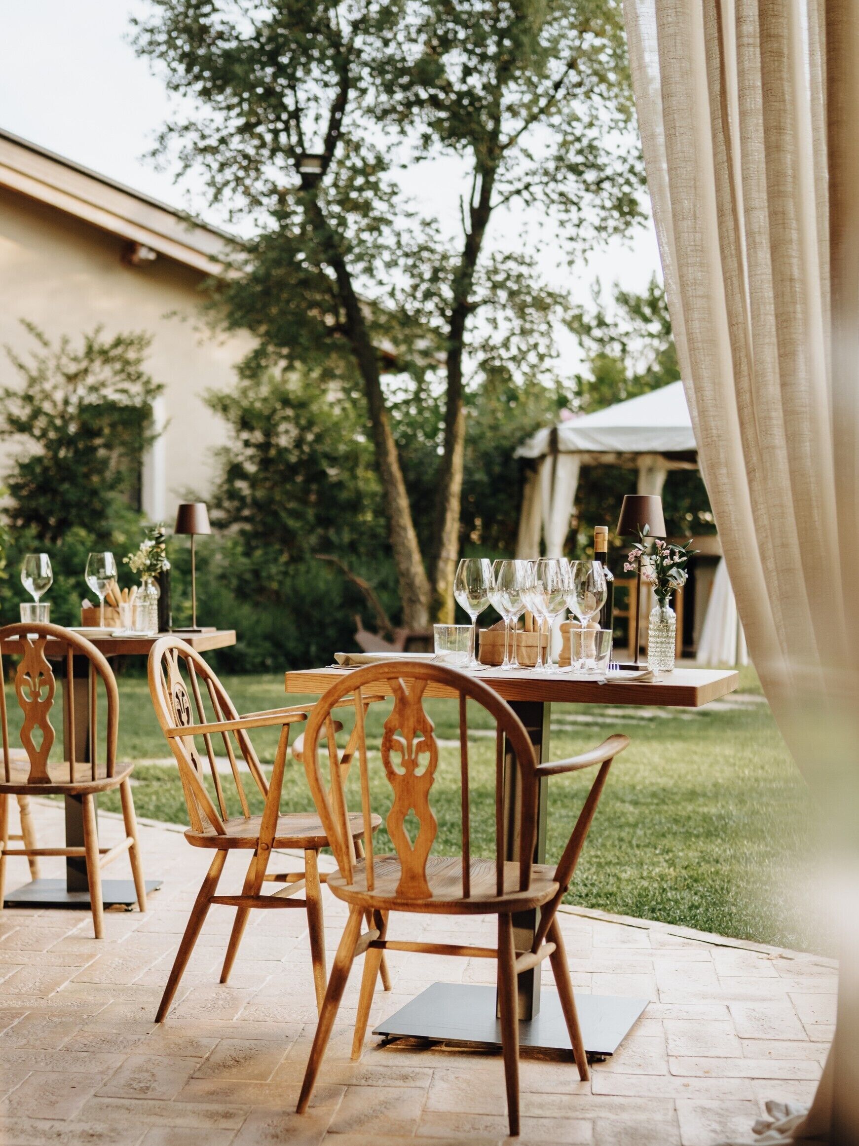 An outdoor dining area with wooden chairs and tables, set against a backdrop of trees and a building.