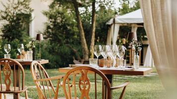 An outdoor dining area with wooden chairs and tables, set against a backdrop of trees and a building.
