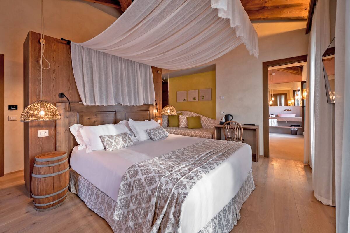A bedroom with a large bed, wooden headboard, and a canopy. There is a nightstand with a lamp and a wooden barrel. The room has wooden flooring and a yellow accent wall.