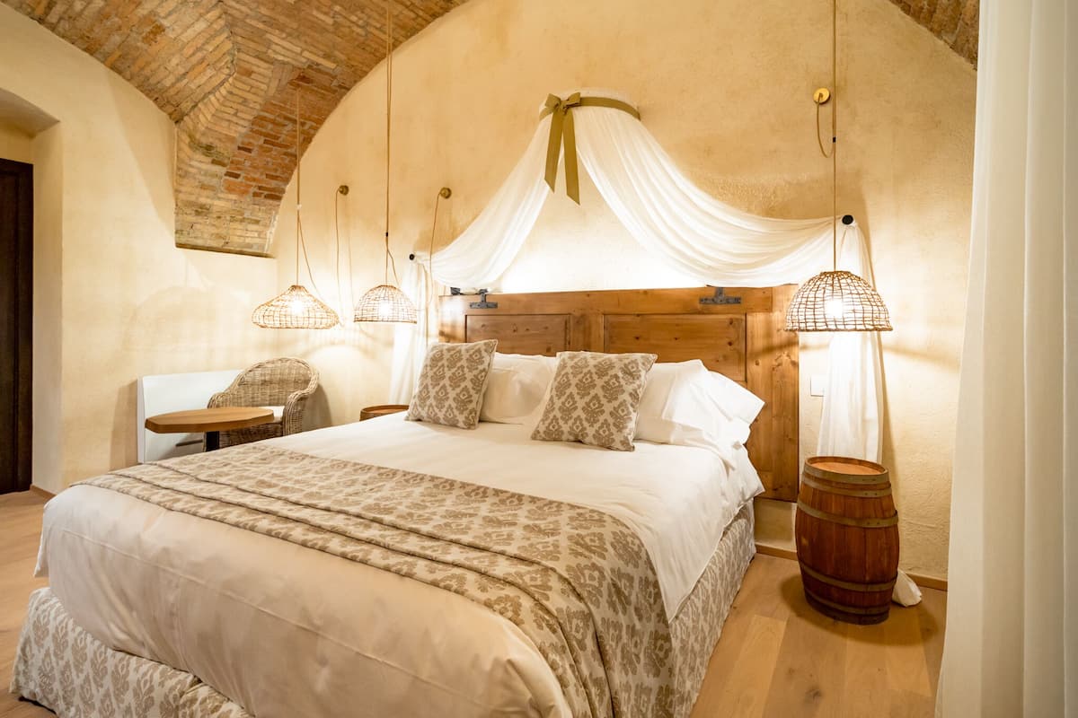 A spacious bedroom with a large bed, a wooden headboard, and a rustic brick ceiling. There are two bedside tables with lamps and a wooden barrel.