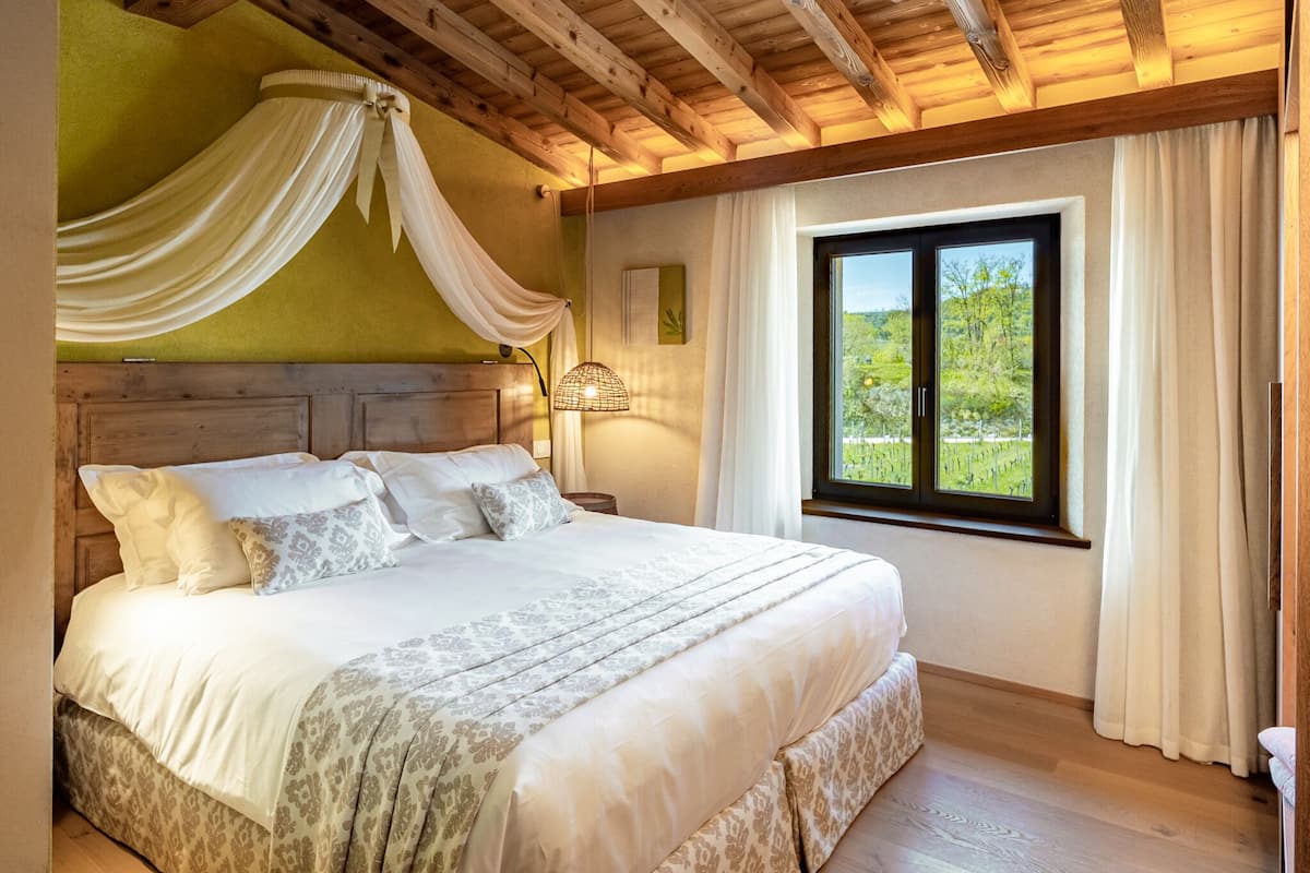 A large bed with a canopy, a wooden headboard, and a view of a green landscape through a window.