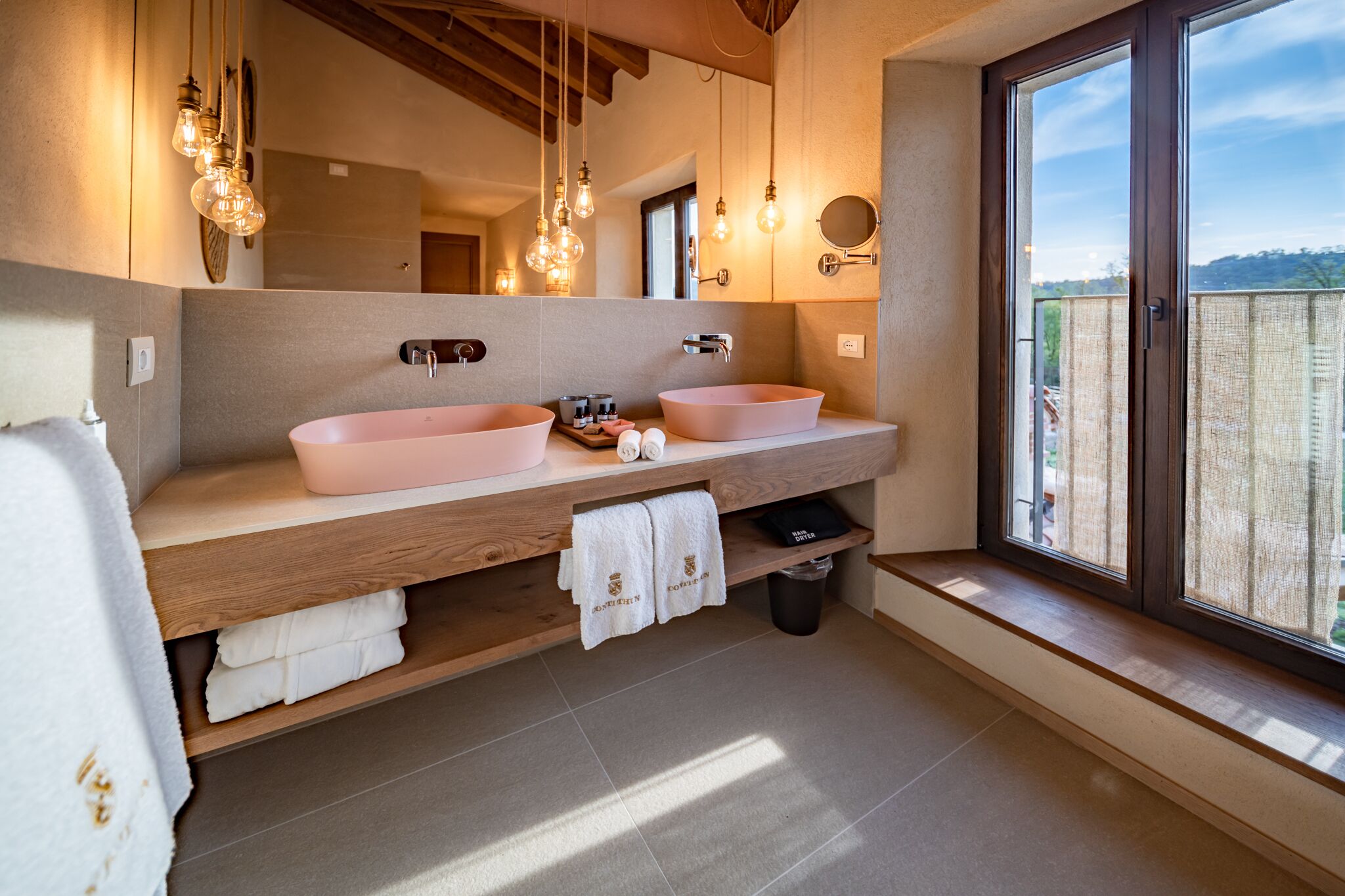 Une salle de bain moderne avec deux lavabos roses, un meuble-lavabo en bois, des luminaires suspendus et une grande fenêtre offrant une belle vue.