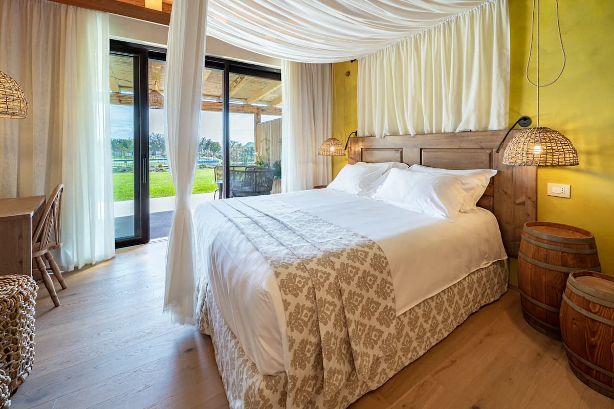 A bedroom with a large bed, a wooden headboard, two bedside lamps, and a view of a garden through a glass door.