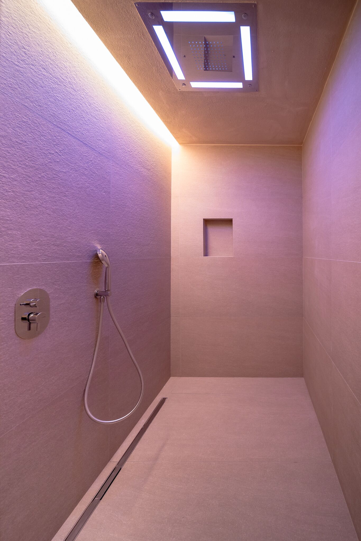 Une douche moderne dotée d’un pommeau de douche à main, d’un luminaire rectangulaire au plafond et d’une petite grille de ventilation carrée sur le mur.