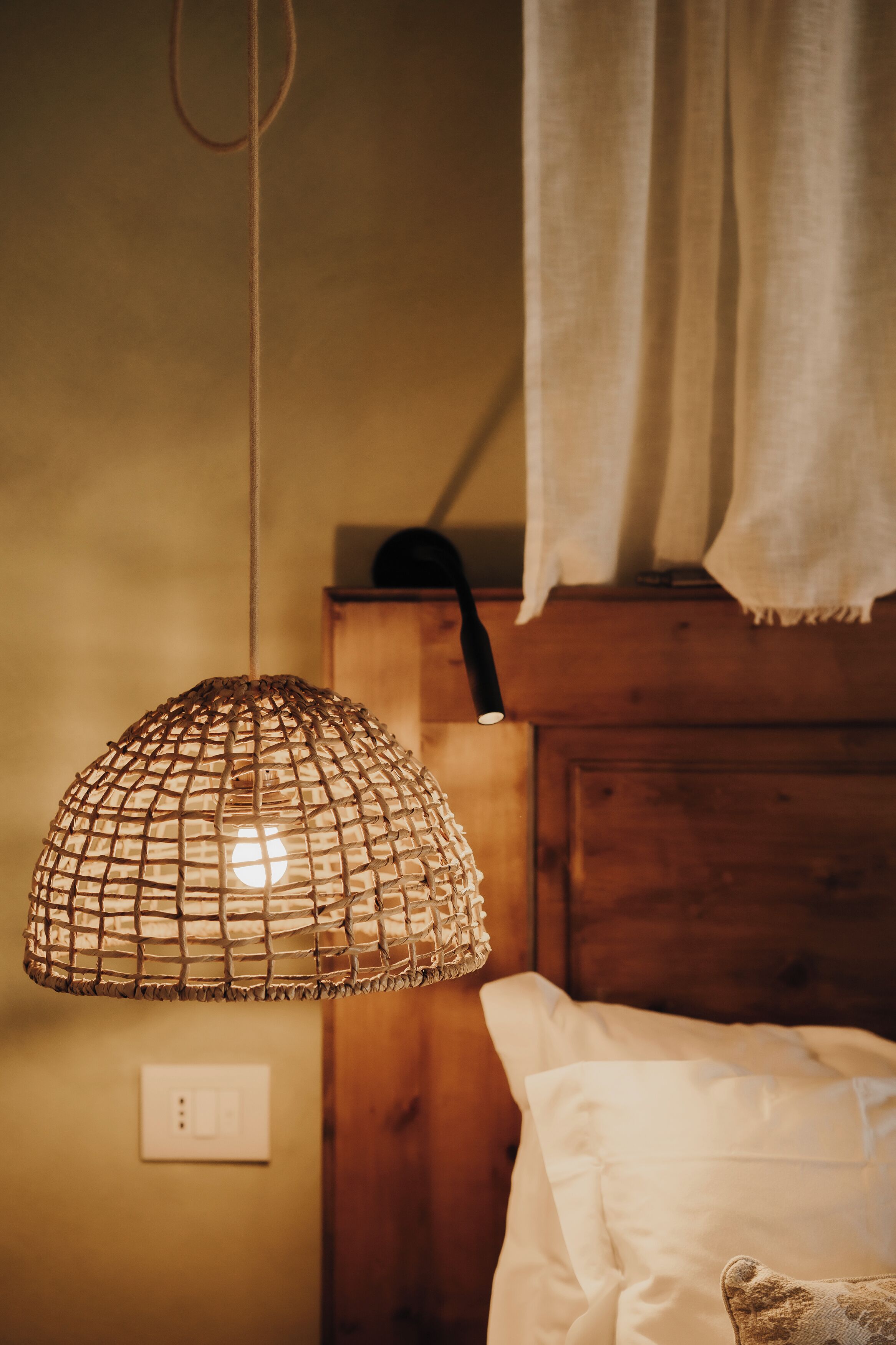 A room with a woven light fixture, wooden headboard, and a white bed.