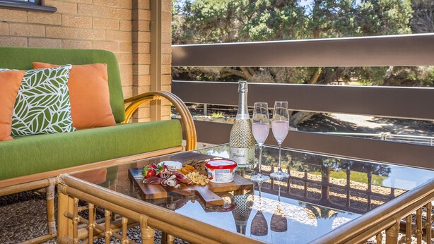 A glass-top table with a bottle, glasses, and food items on a balcony with a view of trees. - Ocean Close - Spacious  Beach House (Guilderton)