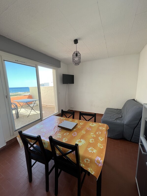 Un coin repas avec une table et des chaises, un téléviseur et une vue sur la plage, accessible par une porte coulissante.