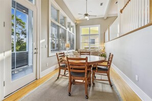 A dining room with a round table and six wooden chairs, large windows with blinds, a ceiling fan, and a view of the outdoors.