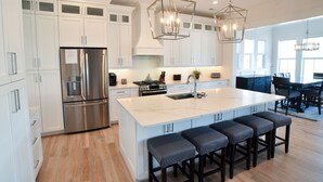 A modern kitchen with white cabinets, a stainless steel refrigerator, a large island with bar stools, and pendant lighting. - New 5 bedroom house in Bayside Village by the beach! (Cape Charles)