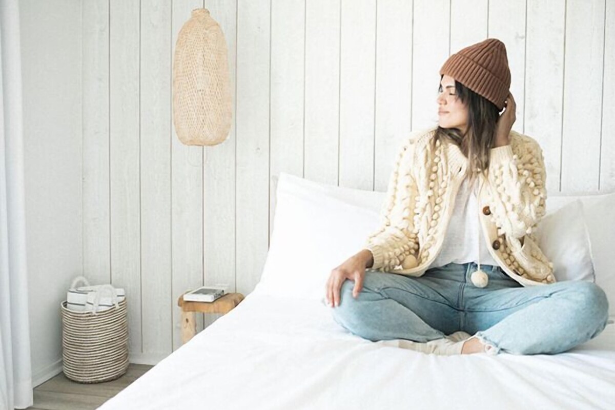 A person sitting on a bed in a room with a white wooden wall, a small wooden nightstand, and a woven basket.