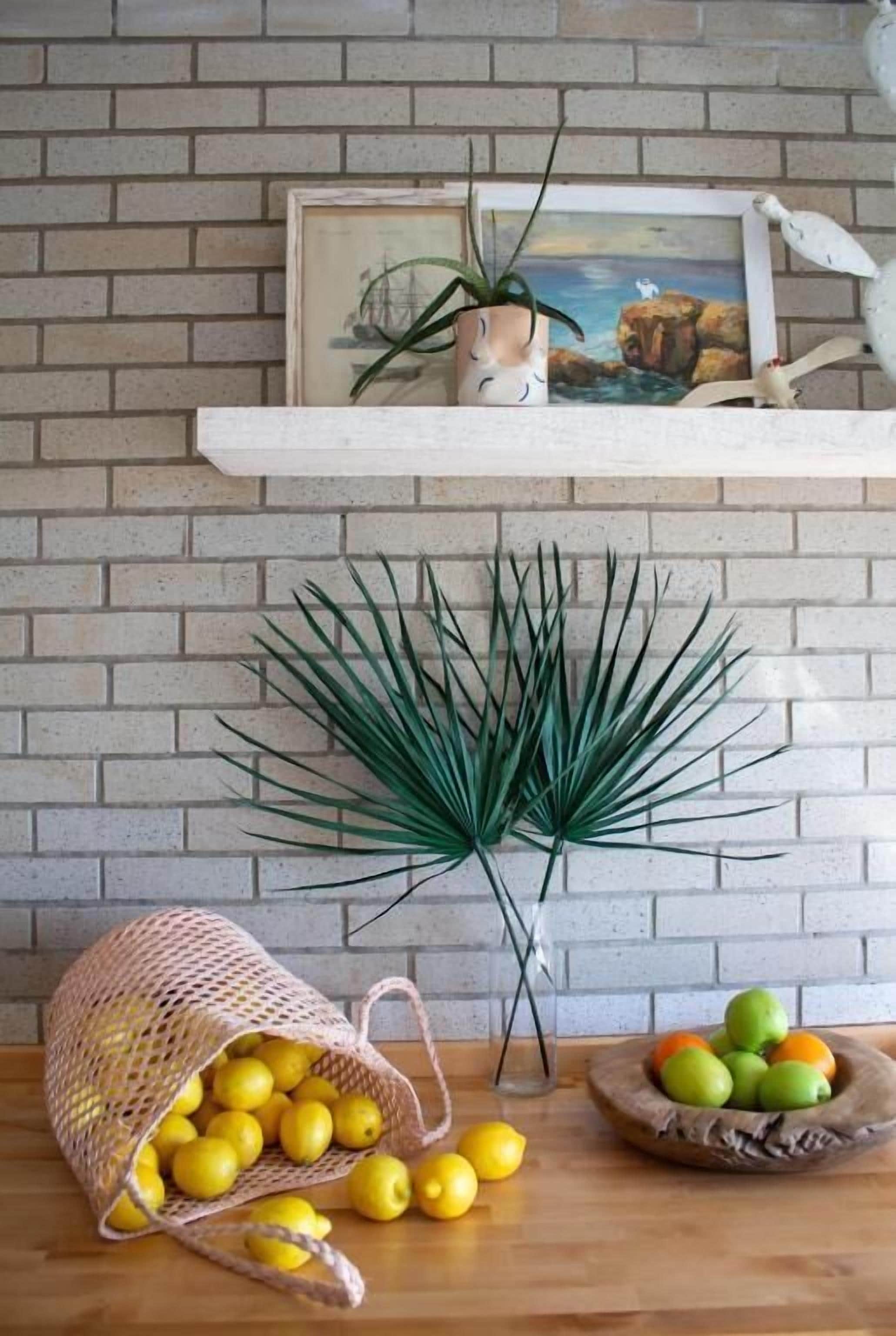 A kitchen with a brick wall, a white shelf with a framed picture, a potted plant, a basket of lemons, and a bowl of green apples.