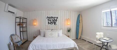 Un dormitorio con una cama, una tabla de surf, una silla, una mesita y una pared con un letrero.