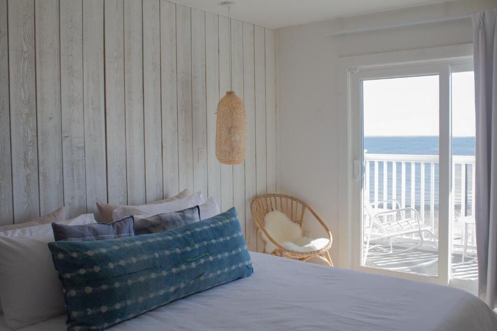 Un dormitorio con cama, almohadas, una silla de mimbre y vista al mar a través de un ventanal corredero.