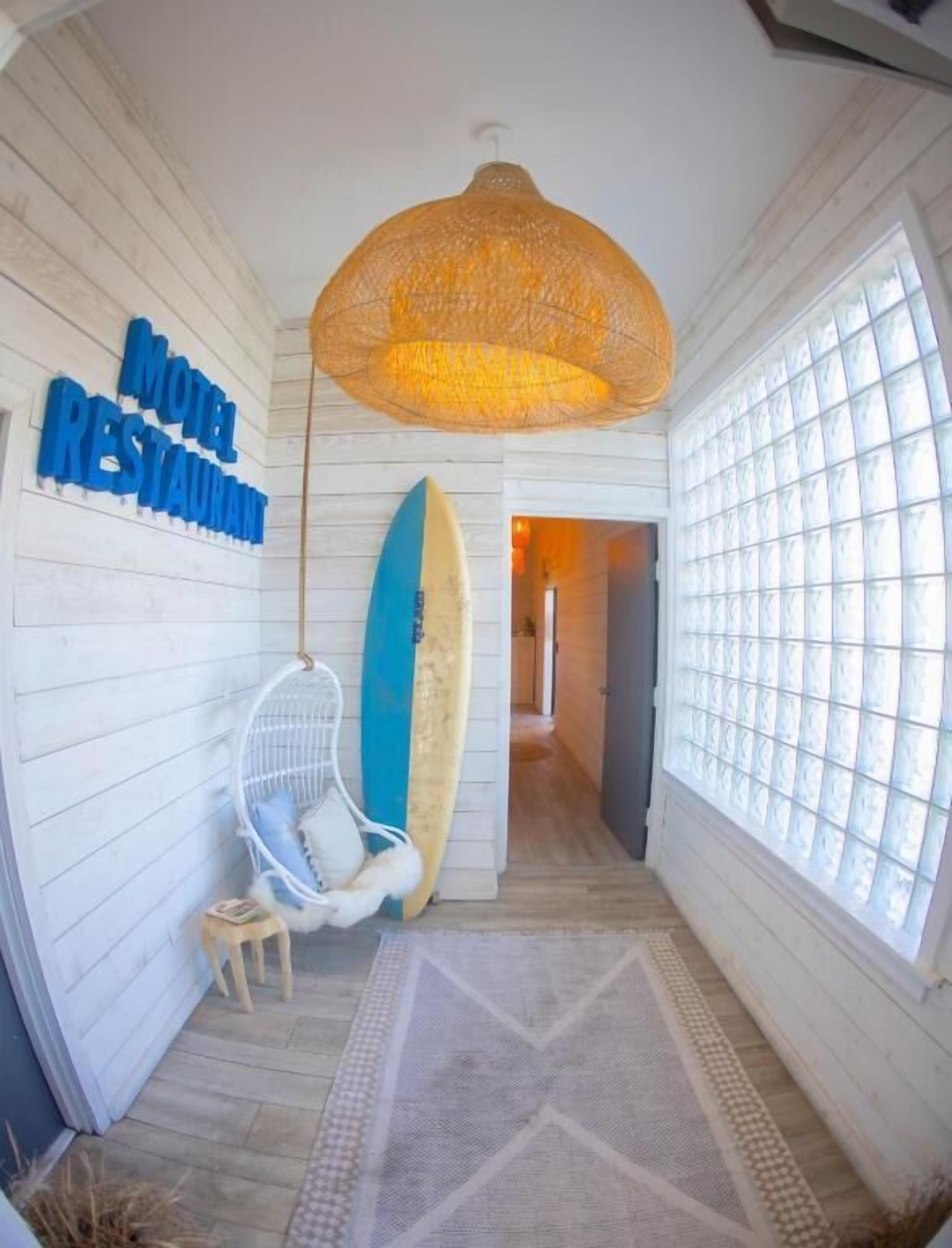 Un pasillo con una lámpara de mimbre colgante, una tabla de surf apoyada contra la pared y un cartel que dice 'Motel Restaurant'.