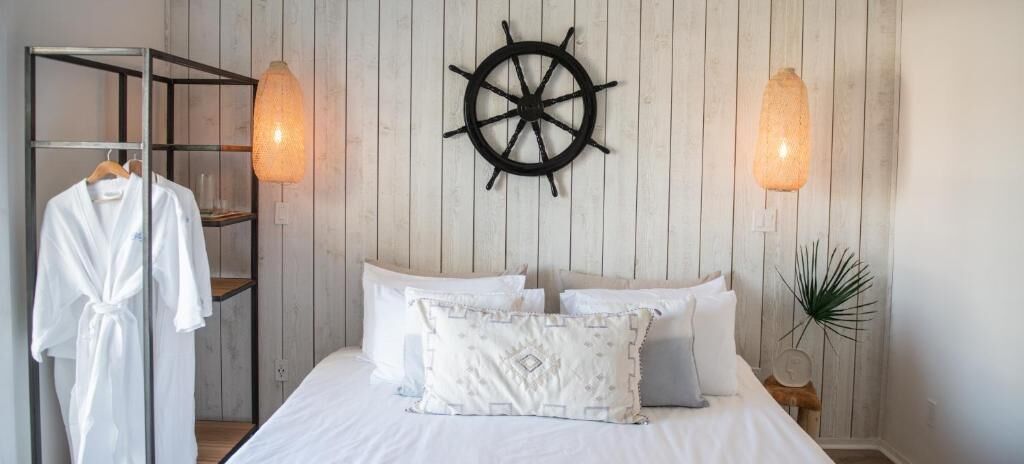 Un dormitorio con pared de madera, una cama con ropa de cama blanca, un adorno con timón de barco y dos lámparas colgantes.