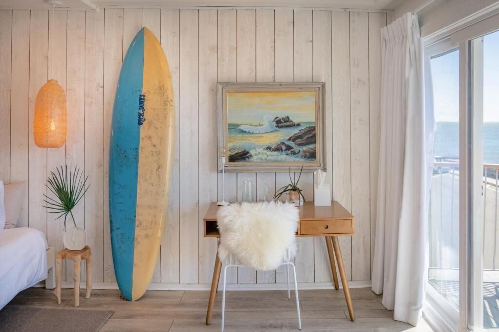 A room with a wooden wall, a surfboard, a painting of a beach, a desk, a chair, and a view of the sea.