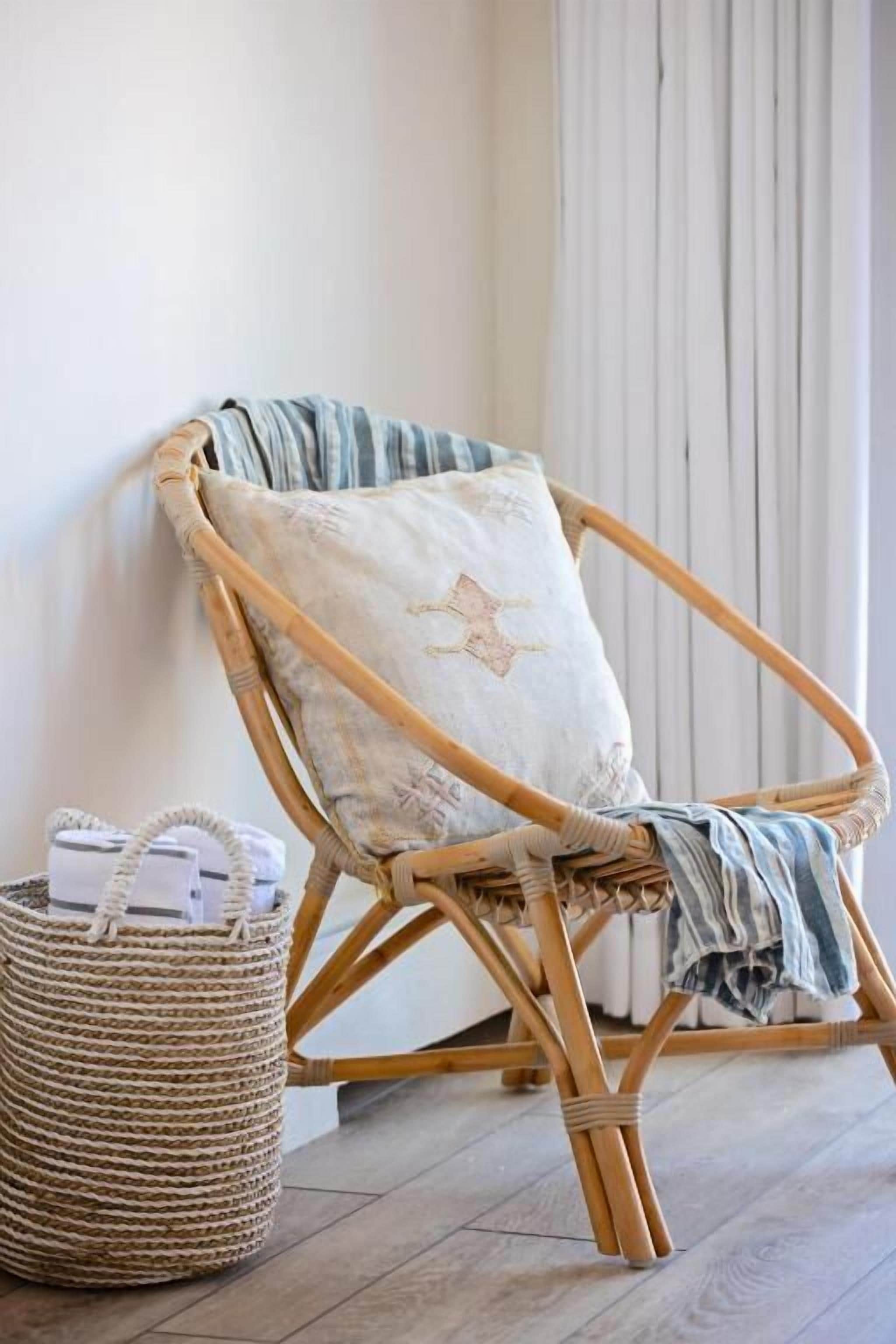 Una silla de mimbre con un cojín apoyada contra una pared, una cesta tejida con objetos, y una manta a rayas sobre el suelo.