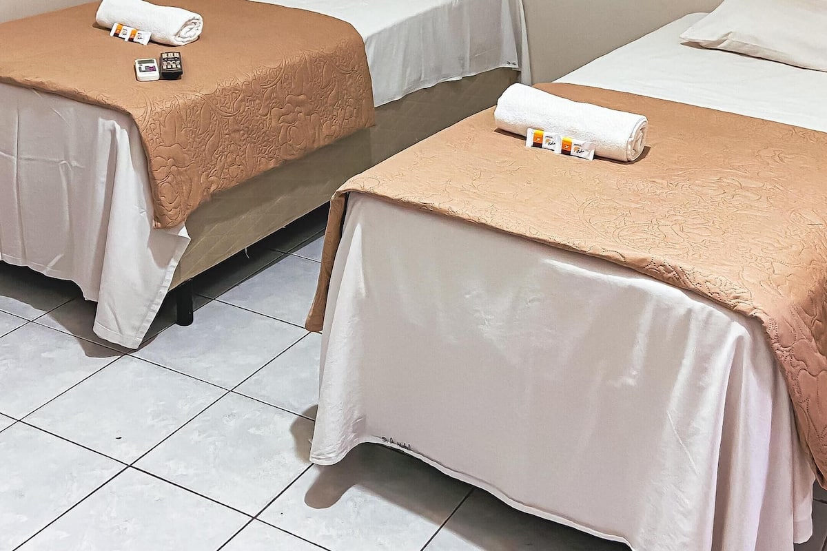 Ett hotellrum med två separata sängar, var och en med ett beige överkast och vita kuddar. Det finns toalettartiklar på sängarna, och rummet har vita kakelplattor på golvet.