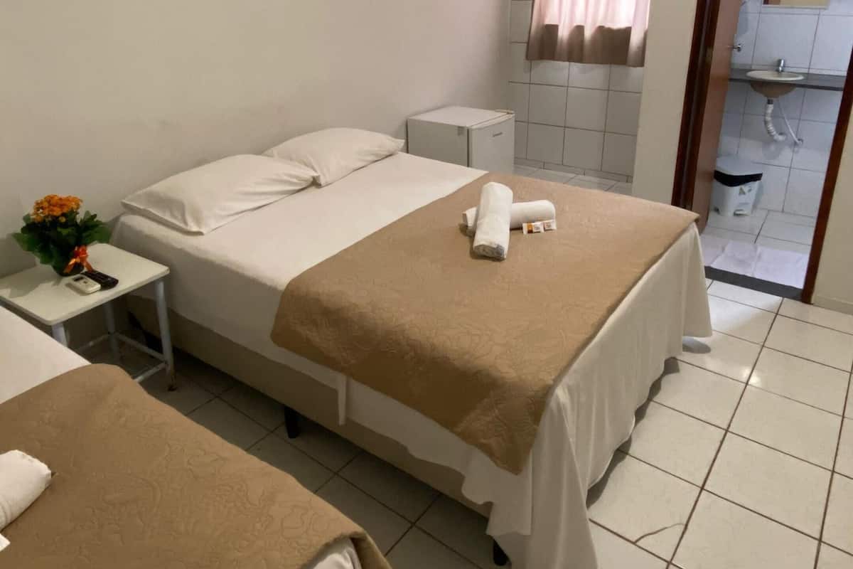 Ett hotellrum med två sängar, ett litet nattduksbord med en vas med blommor, en luftkonditioneringsanläggning, ett fönster med gardiner och ett badrum som syns genom en öppen dörr.