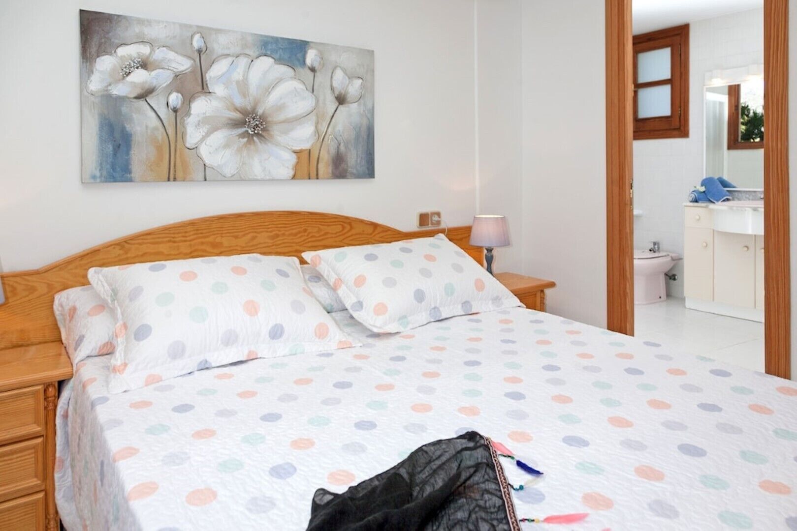 A bedroom with a bed, a wooden headboard, a nightstand, a lamp, and a painting of flowers on the wall.