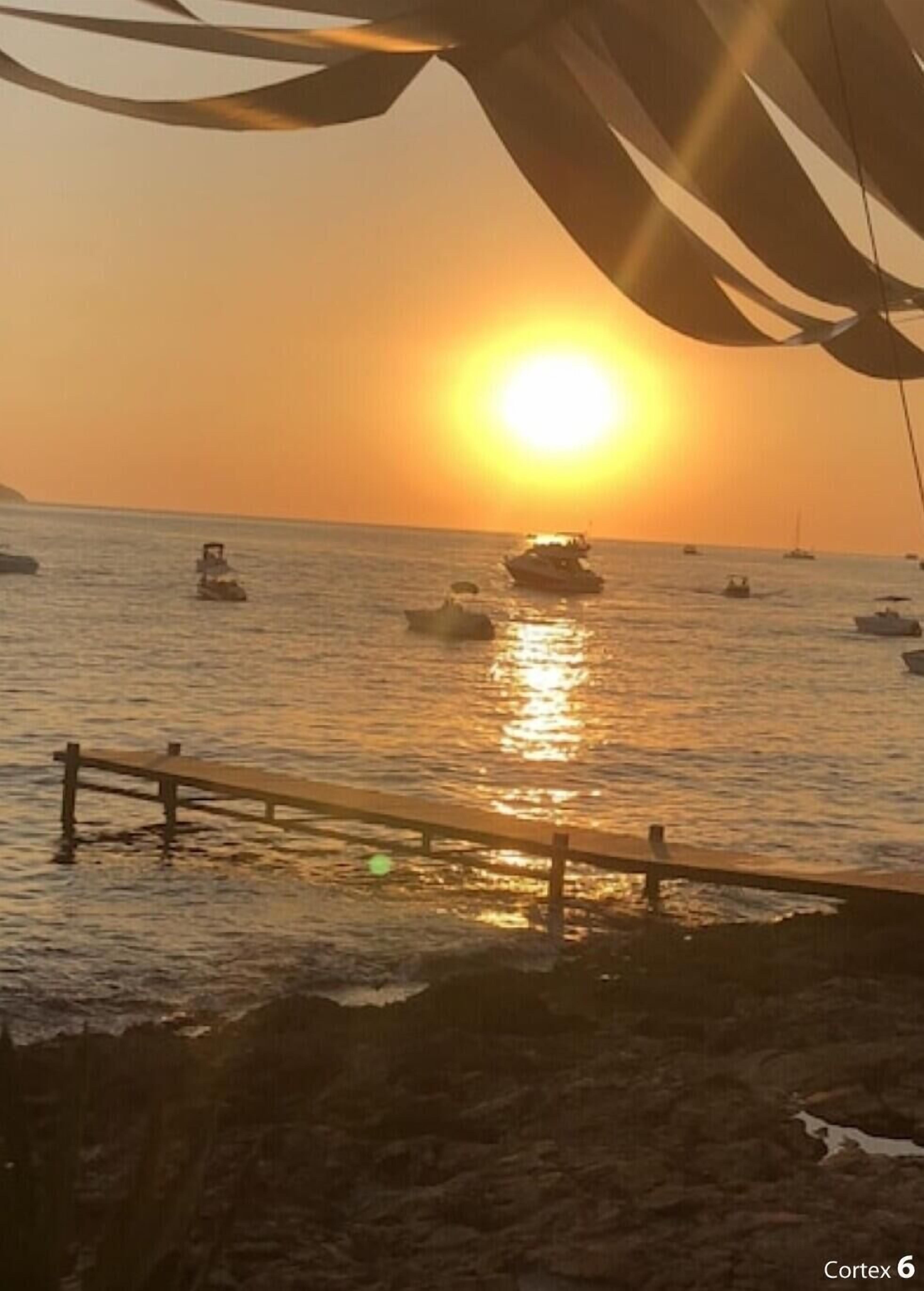 A sunset over a calm sea with boats, a wooden pier, and a rocky shoreline.