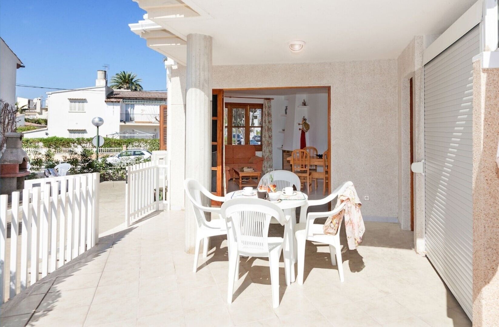 A balcony with a table and chairs, a view of the street, and a building with a closed shutter.