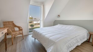 Un dormitorio con una cama grande, una silla de mimbre, una mesita y una ventana con vistas al exterior.