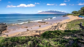 A beach with clear waters, rocky shore, and a mountain in the background. - Hale Kamaole #170, Fully Renovated, Ground Floor, Full A/C, Near Kamaole III (Kihei)