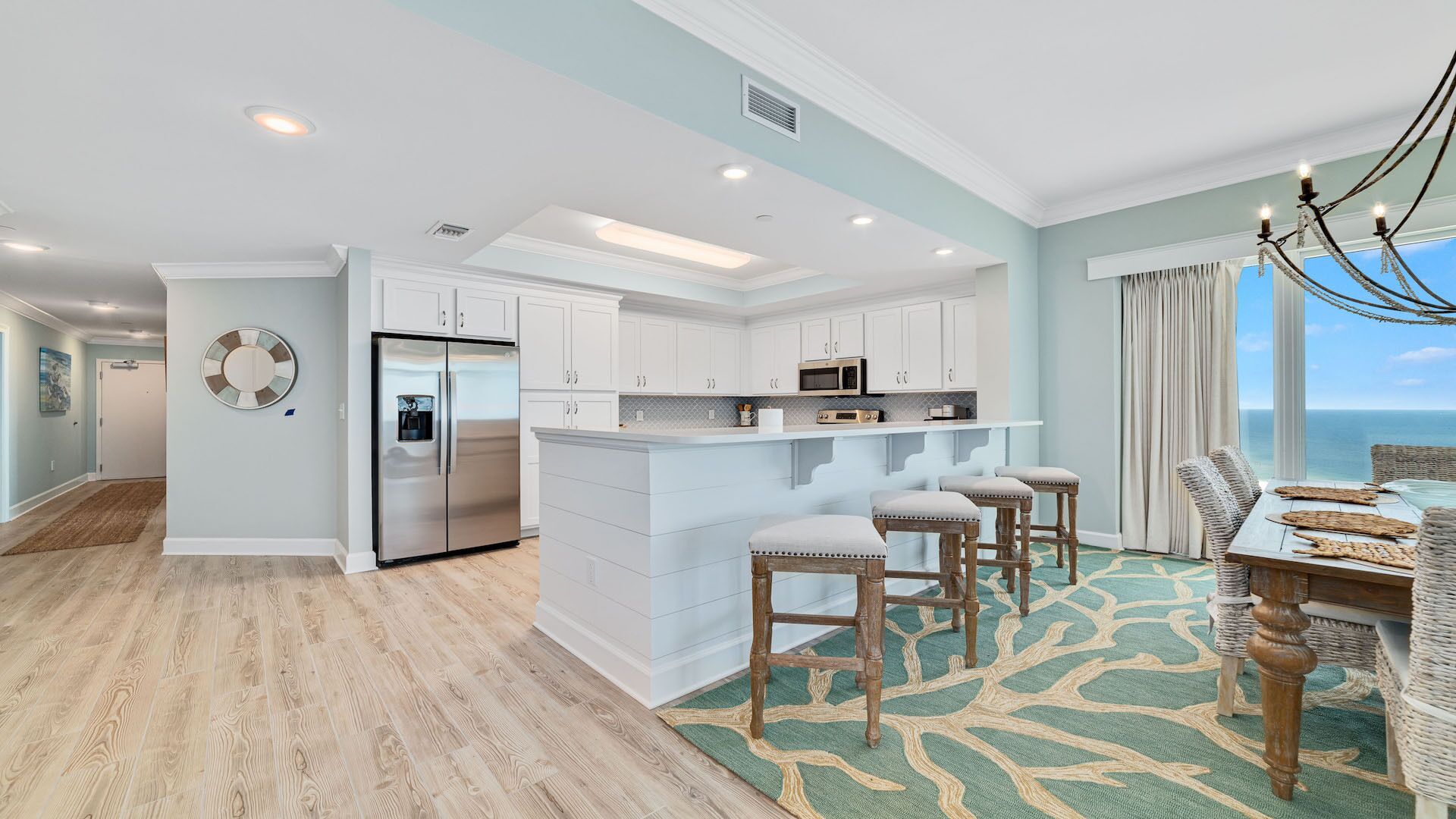 A modern kitchen with white cabinetry, a stainless steel refrigerator, and a dining area with wicker chairs and a view of the ocean.