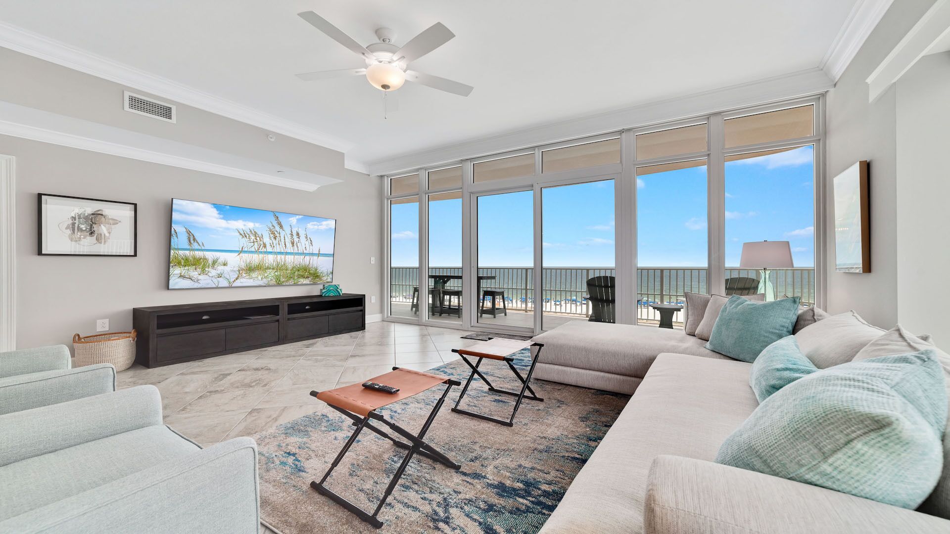 A spacious living room with a large window overlooking the beach, a ceiling fan, and a flat-screen TV mounted on the wall.