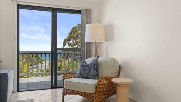 Una silla de mimbre con cojines, una mesita redonda pequeña y una lámpara de pie en una habitación con vistas al mar a través de un ventanal corredero.