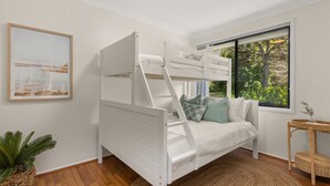 Una cama litera con escalera, un sofá con cojines, una mesita de mimbre, un cuadro enmarco y una planta en maceta.