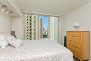 Une chambre avec un grand lit, une commode en bois et une vue sur de hauts immeubles, visible à travers une porte coulissante.