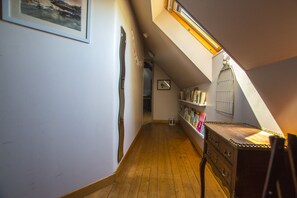 Ein schmaler Flur mit Holzboden, einer geneigten Decke mit Dachfenster, einem Bücherregal und einem kleinen weißen Stuhl.