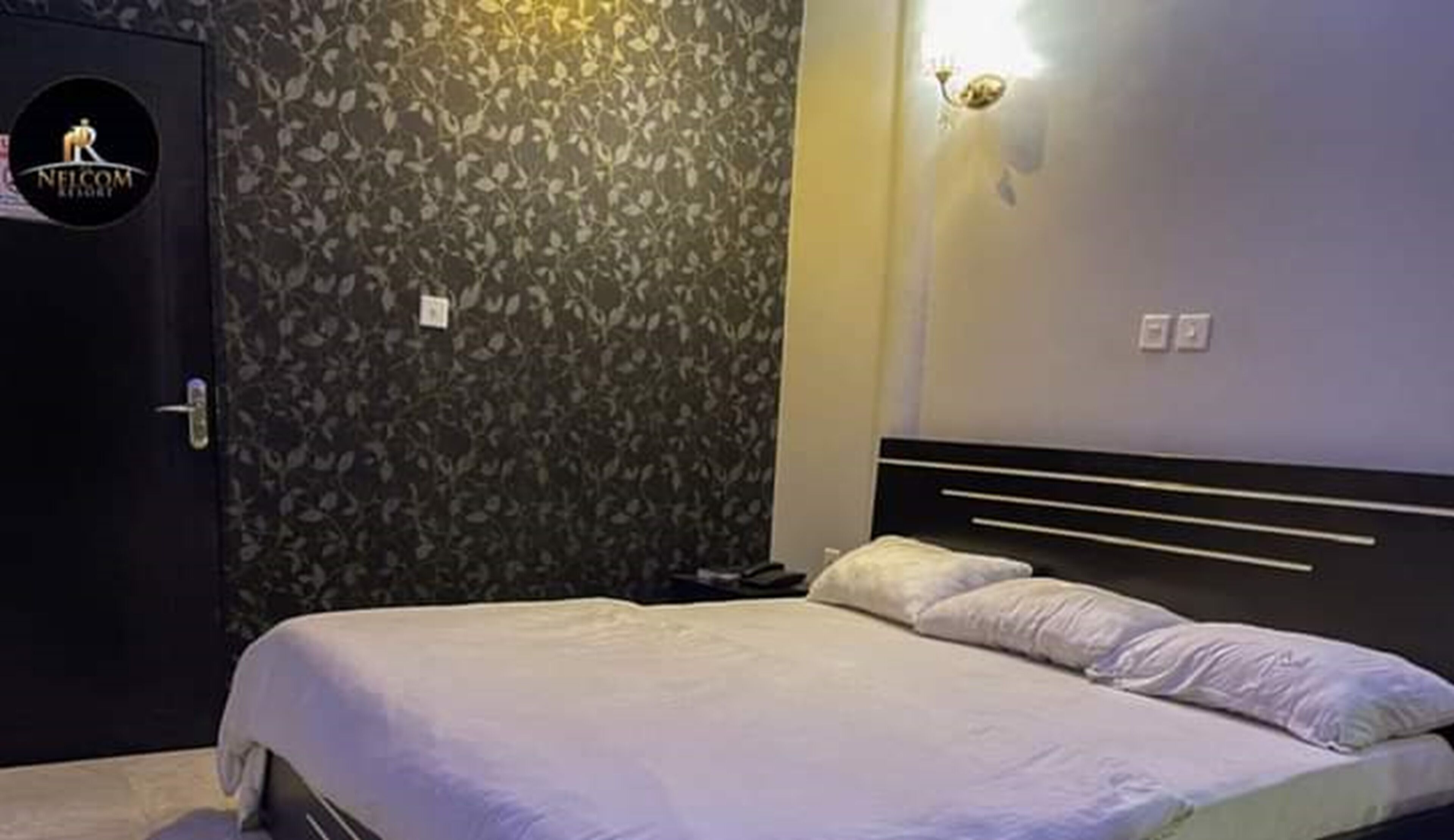Quarto de hotel com cama, mesa de cabeceira, relógio na parede e porta com logotipo.