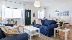 A modern living room with a blue sofa, white coffee table, and a dining area with white chairs and a table. - Villa som das Ondas I (Budens)