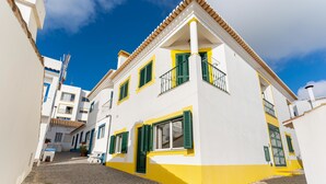 A two-story building with a white facade and yellow accents, featuring a cobblestone street and a clear blue sky. - Villa som das Ondas I (Budens)