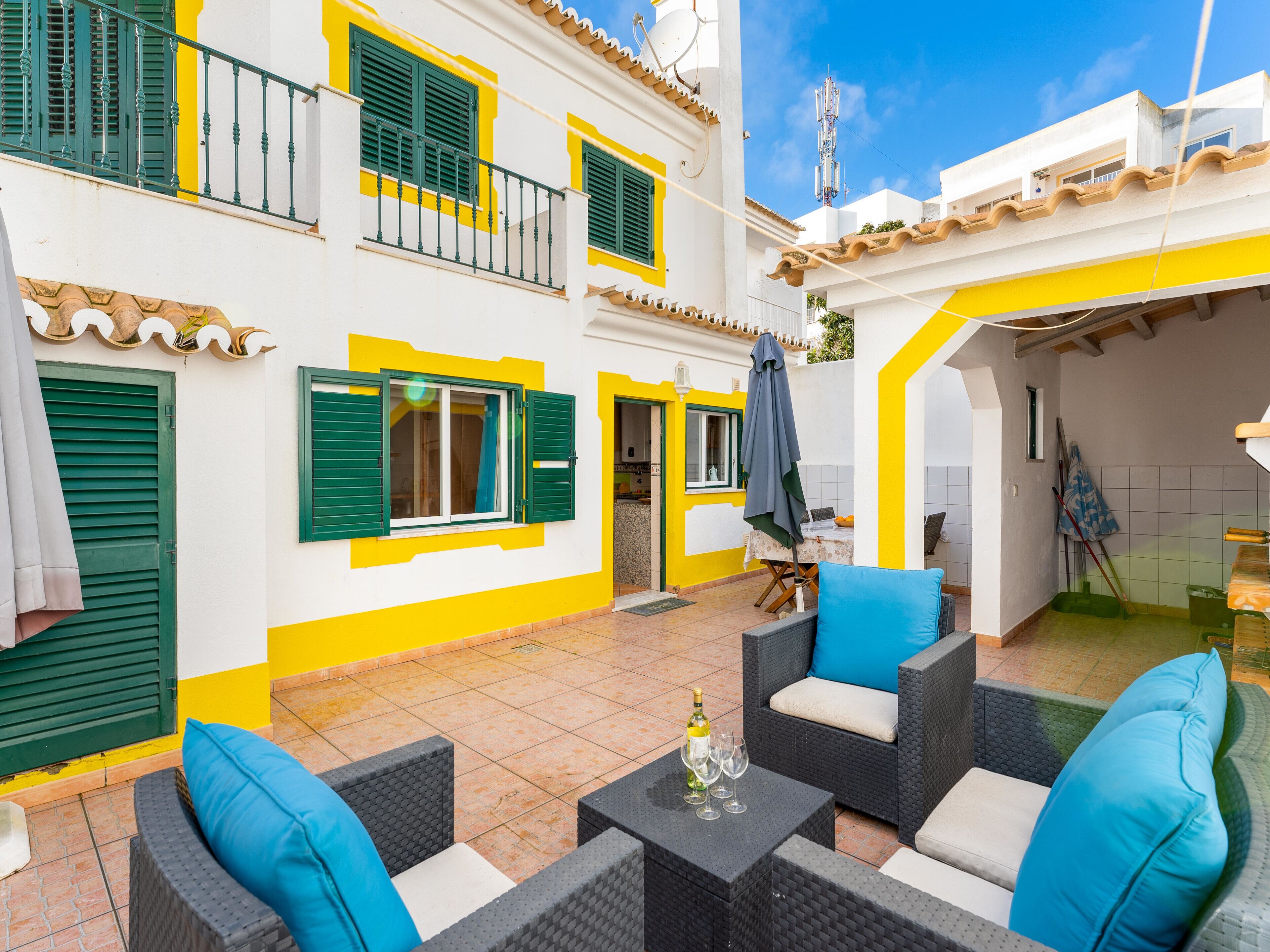 A courtyard with outdoor seating, a tiled floor, and a building with yellow and white walls and green shutters.