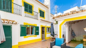 A courtyard with outdoor seating, a tiled floor, and a building with yellow and white walls and green shutters. - Villa som das Ondas I (Budens)
