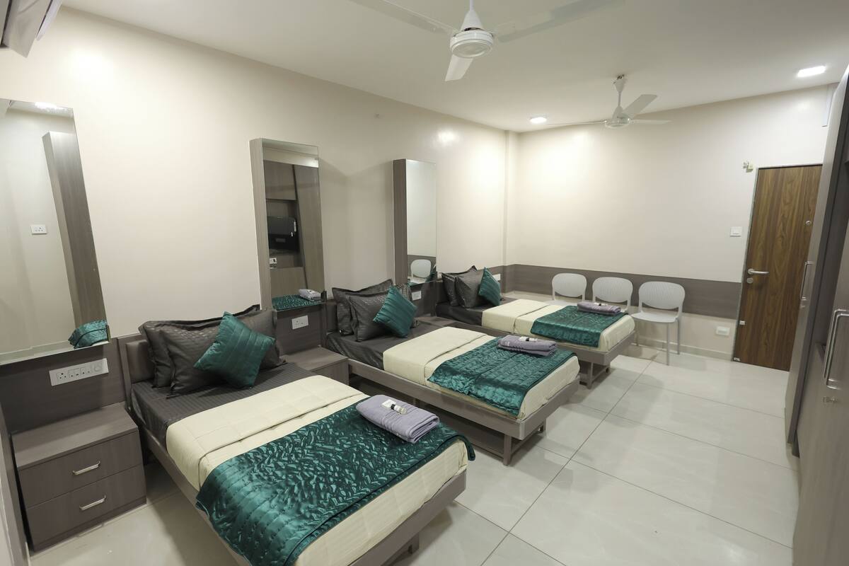 A hotel room with three beds, each with a green bedspread, a bedside table, and a mirror. There are ceiling fans and a door visible.