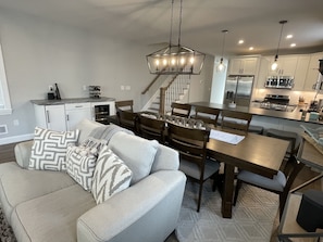 A modern kitchen with white cabinets, a dining area with a wooden table and chairs, and a gray sofa with patterned pillows.